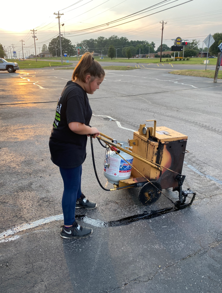 A person uses a walk-behind asphalt crack sealing machine to repair a pavement surface in a parking lot at sunset.