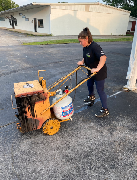 A person pushes a yellow asphalt melter on wheels with a propane tank attached, painting a line in a parking lot.