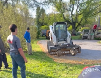 A Bobcat skid-steer loader sits on a gravel drive with people standing nearby on a grassy lawn in a wooded setting.