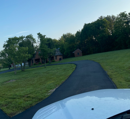 A view from a vehicle looking down a paved, curved driveway toward a brick house and a small shed on a grassy lawn.