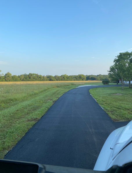 A freshly paved black asphalt driveway winds through a grassy field under a clear blue sky.