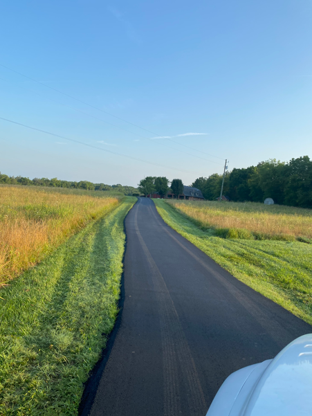 A paved road stretches through a rural landscape with grassy fields on either side under a clear blue sky.