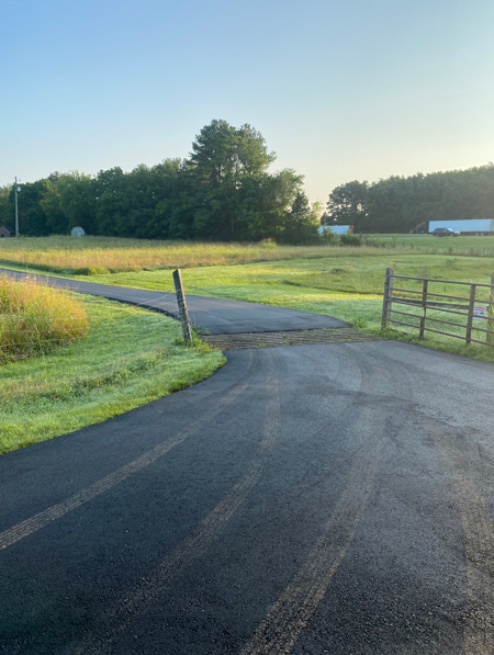 A paved driveway leads into a rural field with a wooden fence on the right under a clear, bright morning sky.