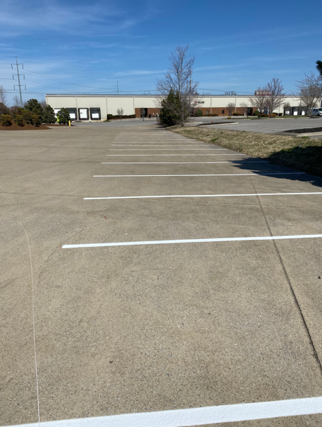 A row of empty parking spaces on a concrete lot in front of a commercial warehouse building under a clear blue sky.