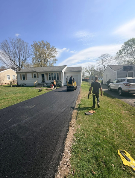 Workers pave a new asphalt driveway leading to a suburban house under a clear blue sky.