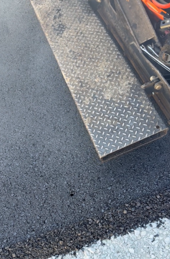 A rectangular, metallic, diamond-plate step extends over a freshly paved black asphalt surface.