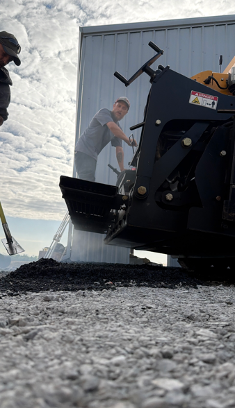 Two people operate heavy machinery to spread asphalt on a gravel ground next to a white metal building.