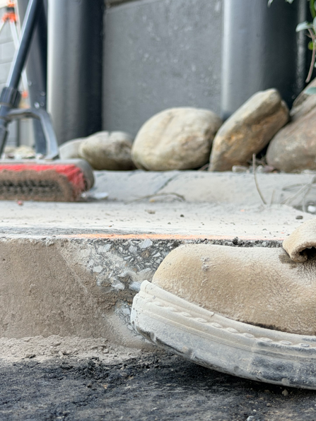 A work boot rests on a concrete step near construction debris and smooth stones.