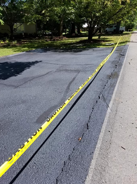 A freshly seal-coated asphalt driveway blocked by yellow caution tape alongside a paved road.