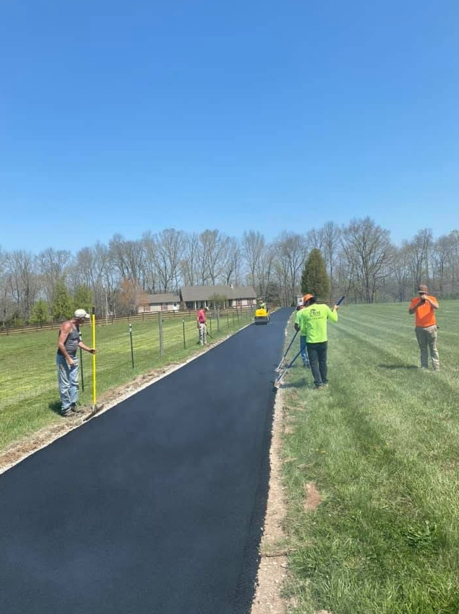 A crew lays fresh black asphalt for a new driveway on a sunny day in a grassy rural area.