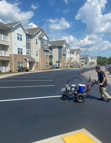 A worker uses a walk-behind machine to paint white parking space lines on a fresh asphalt lot by an apartment complex.