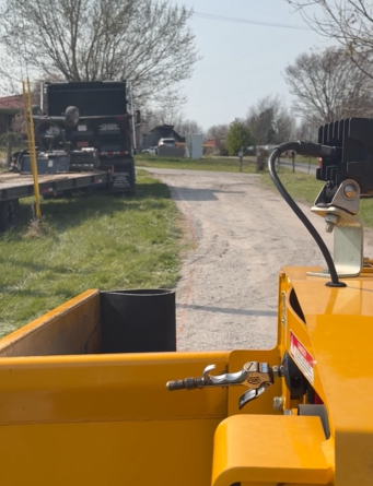 View from a yellow piece of construction machinery showing a gravel path and a flatbed trailer in a grassy field.