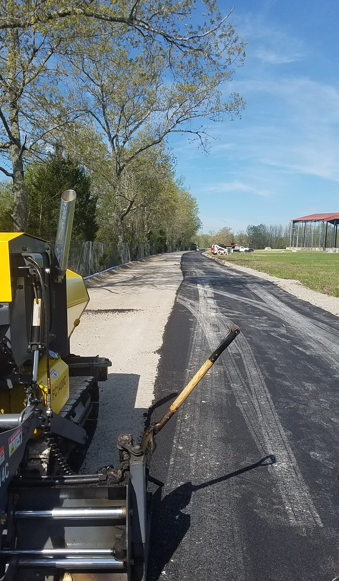 A view from a yellow asphalt paver showing a newly laid blacktop path alongside trees and a grassy field on a sunny day.