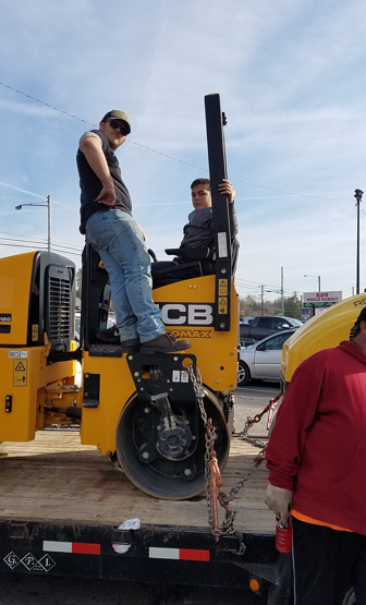 Two people on a yellow JCB road roller parked on a trailer in an outdoor lot.
