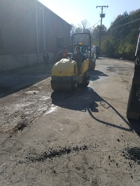 A yellow construction roller drives on an asphalt surface near a building under a bright, sunny sky.