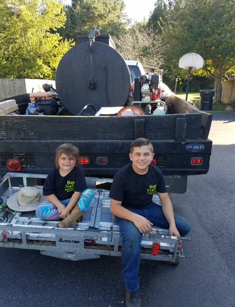 Two smiling people sitting on the back of a black utility truck parked on an asphalt surface, outdoors in daylight.