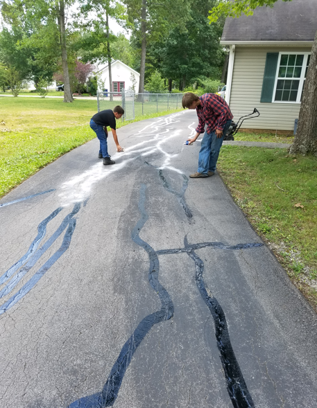 Two people seal asphalt cracks on a residential driveway using a black sealant and white powder.
