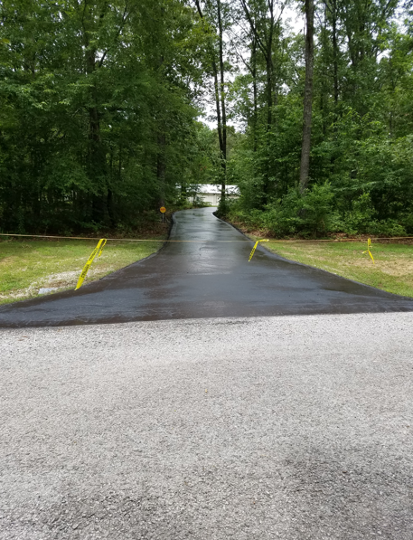 A newly paved asphalt driveway leads into a wooded area, blocked by yellow caution tape.