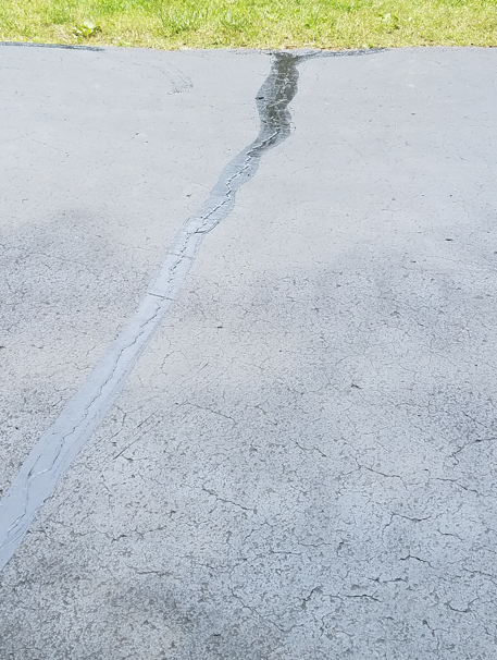 A long, jagged crack running through a gray asphalt driveway near a patch of green grass.