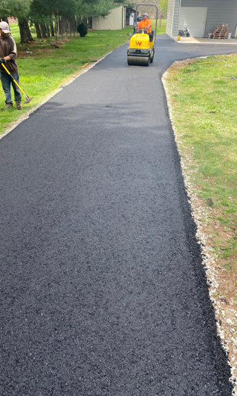 A worker finishes the edges of a freshly paved blacktop driveway as a yellow steamroller travels in the distance.