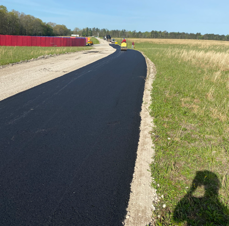 A freshly paved black asphalt road extends through a grassy field under a clear blue sky, with construction in progress.