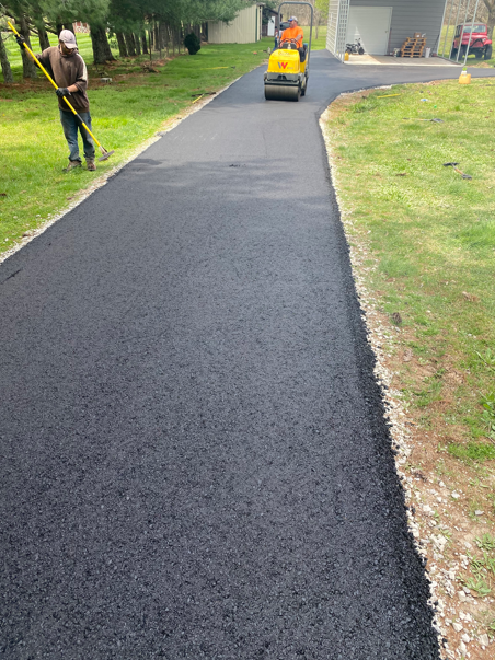 A worker uses a rake on the edge of a newly paved asphalt driveway while a person operates a yellow steamroller ahead.