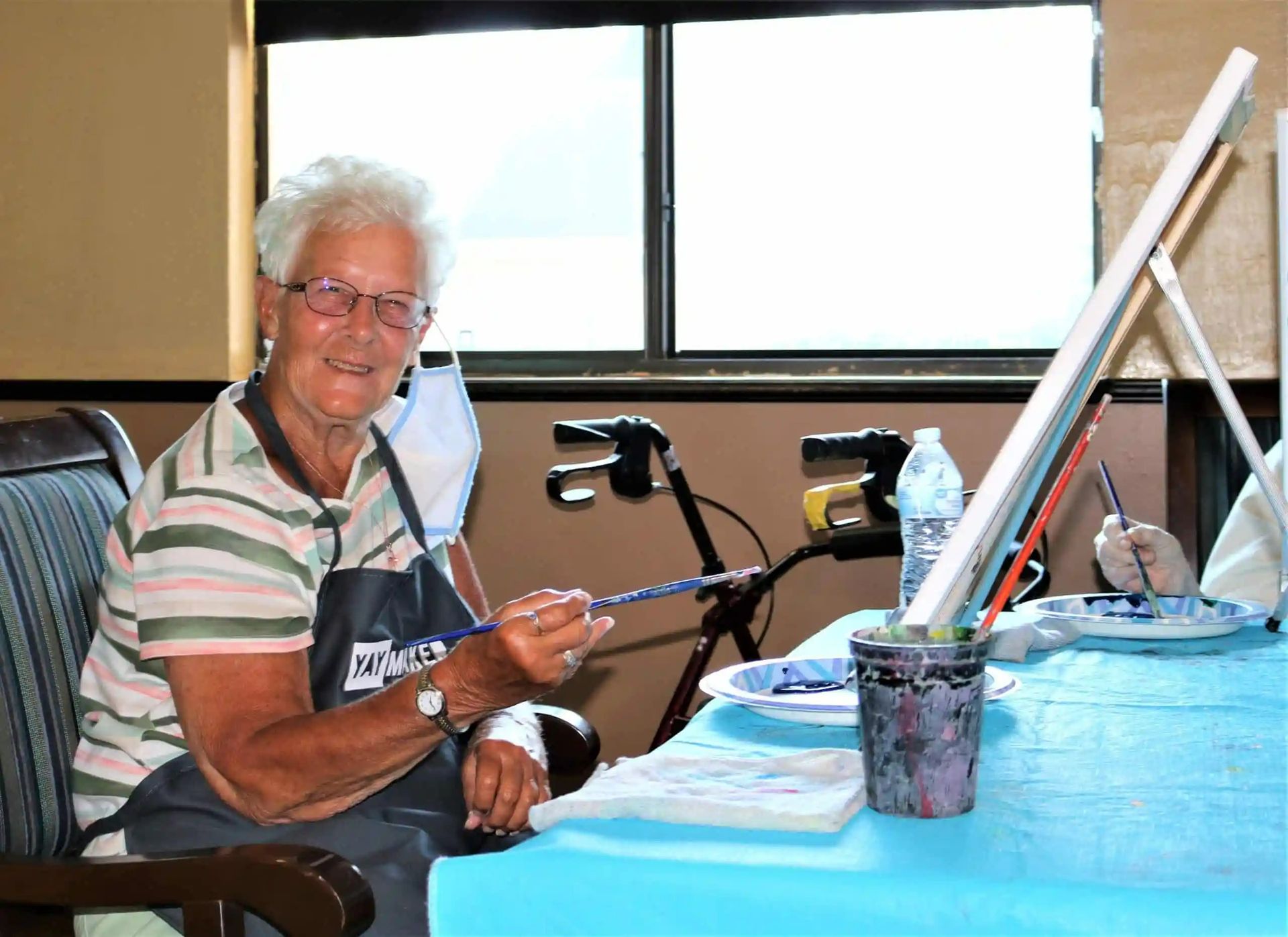 Woman painting at a table, smiling at the camera, with a walker nearby.