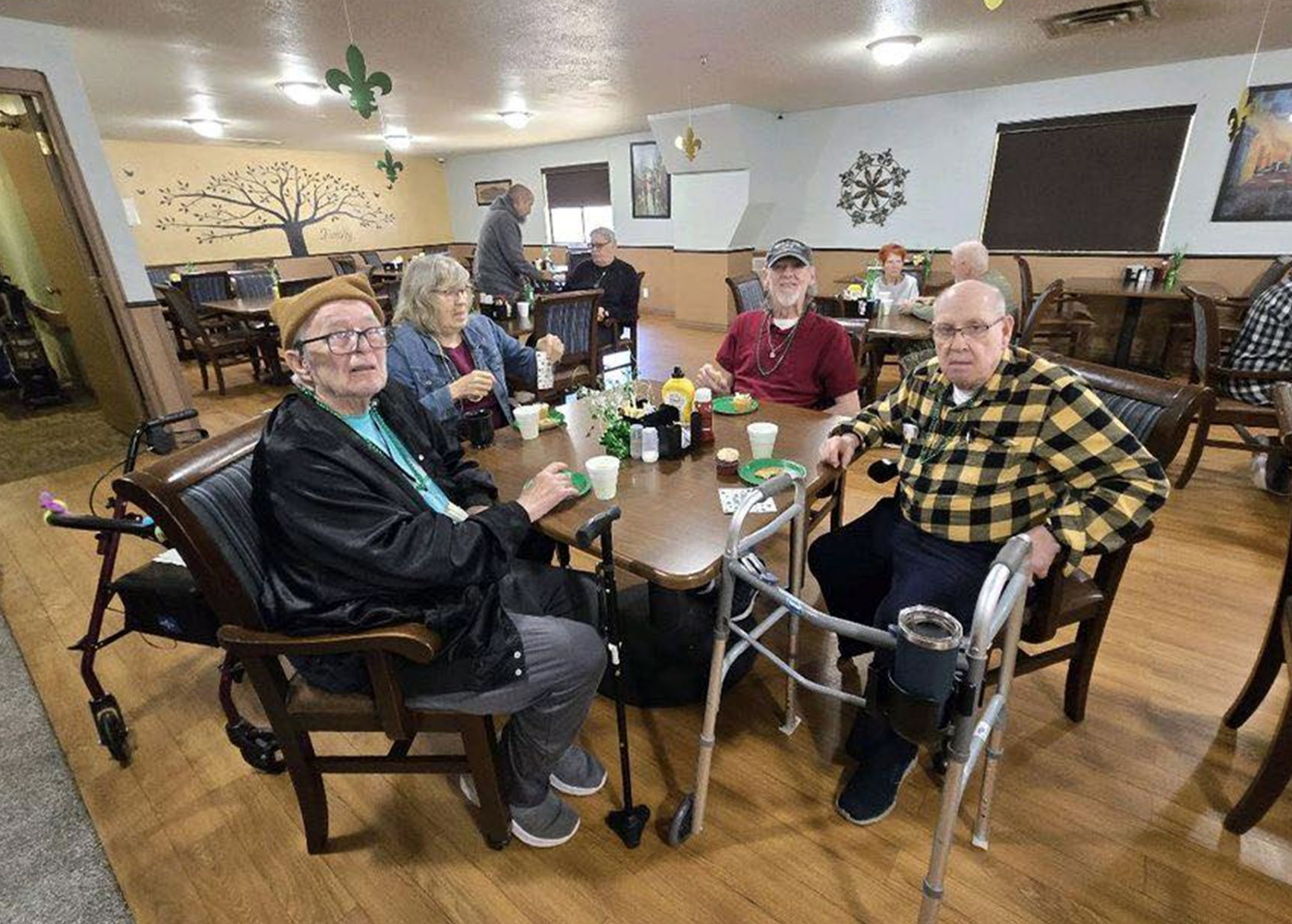 Seniors at a table in a dining room, some using walkers, smiling, decorated for a celebration.