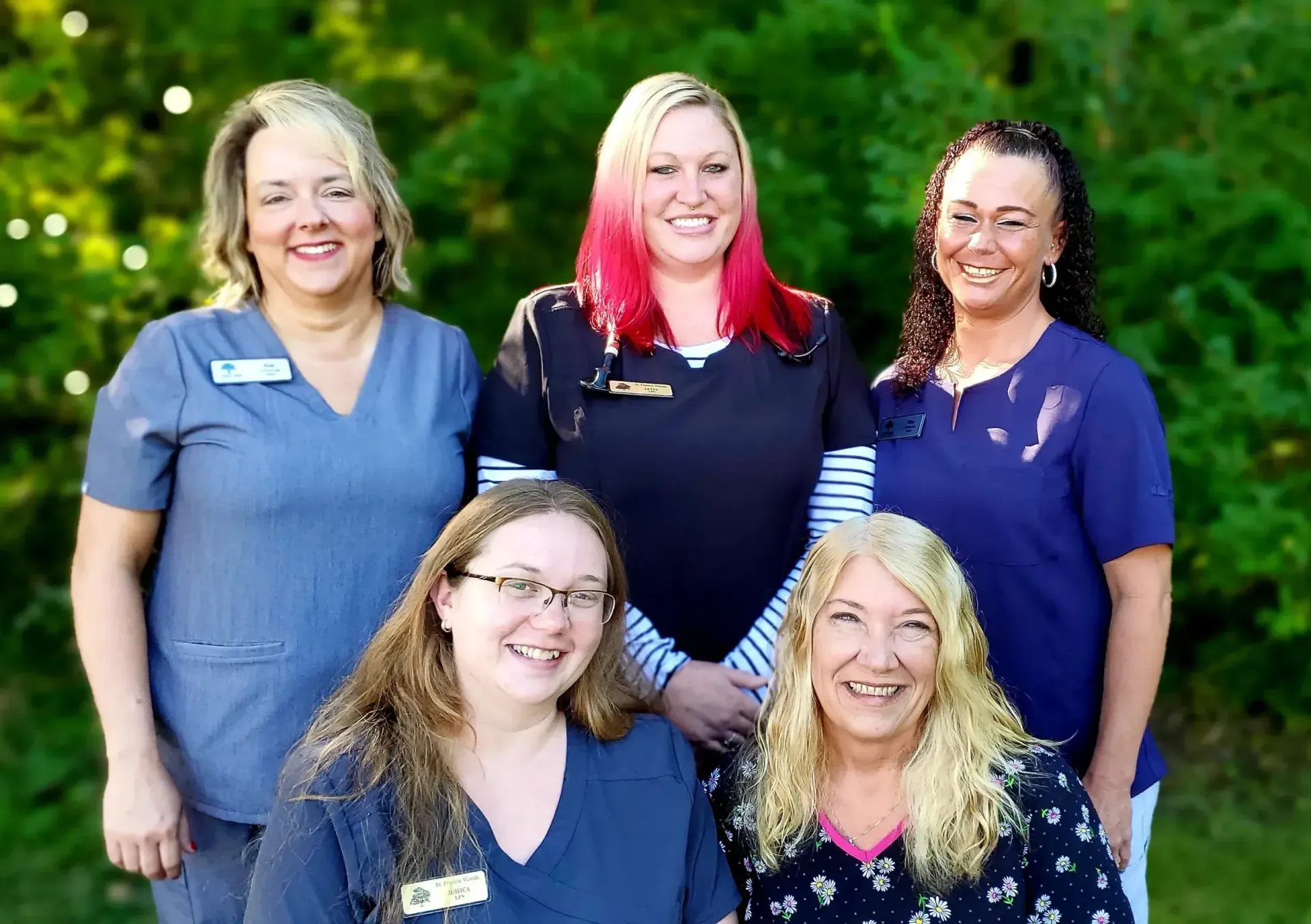 Five women in scrubs smiling outdoors, posing for a group photo.