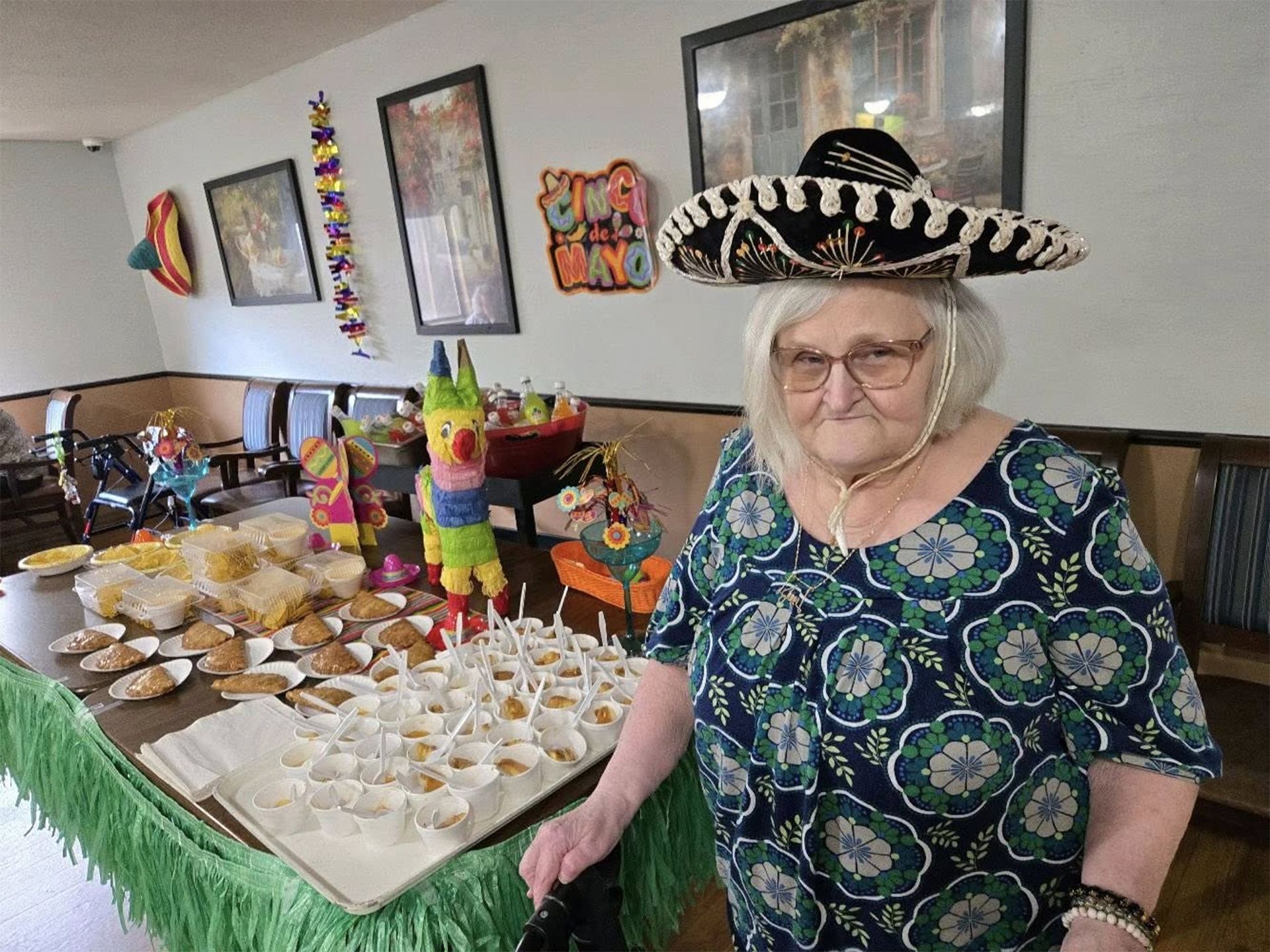 Woman in sombrero stands by a table of food with decorations, presumably at a Cinco de Mayo celebration.