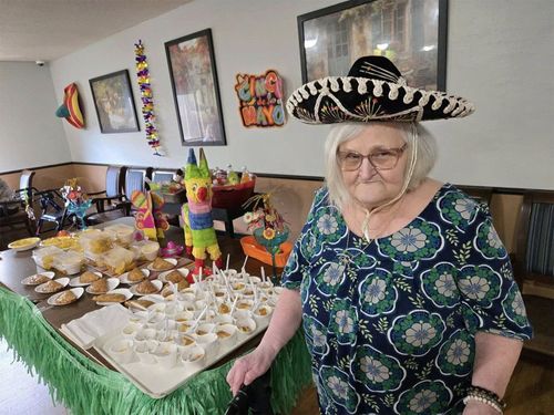 Woman in sombrero stands by a table of food with decorations, presumably at a Cinco de Mayo celebration.