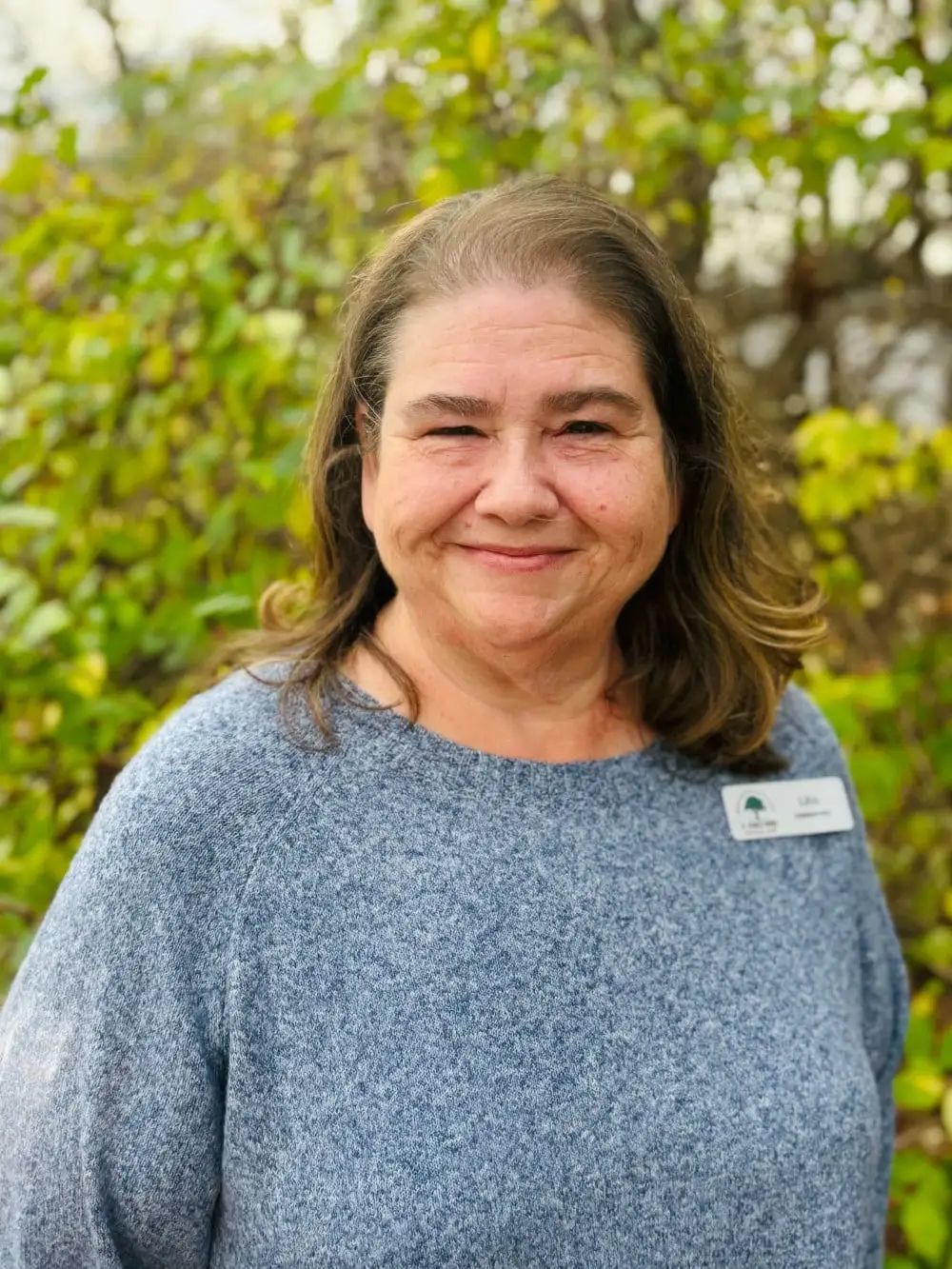 Woman with brown hair in a blue sweater smiles, standing outdoors with green foliage in the background.