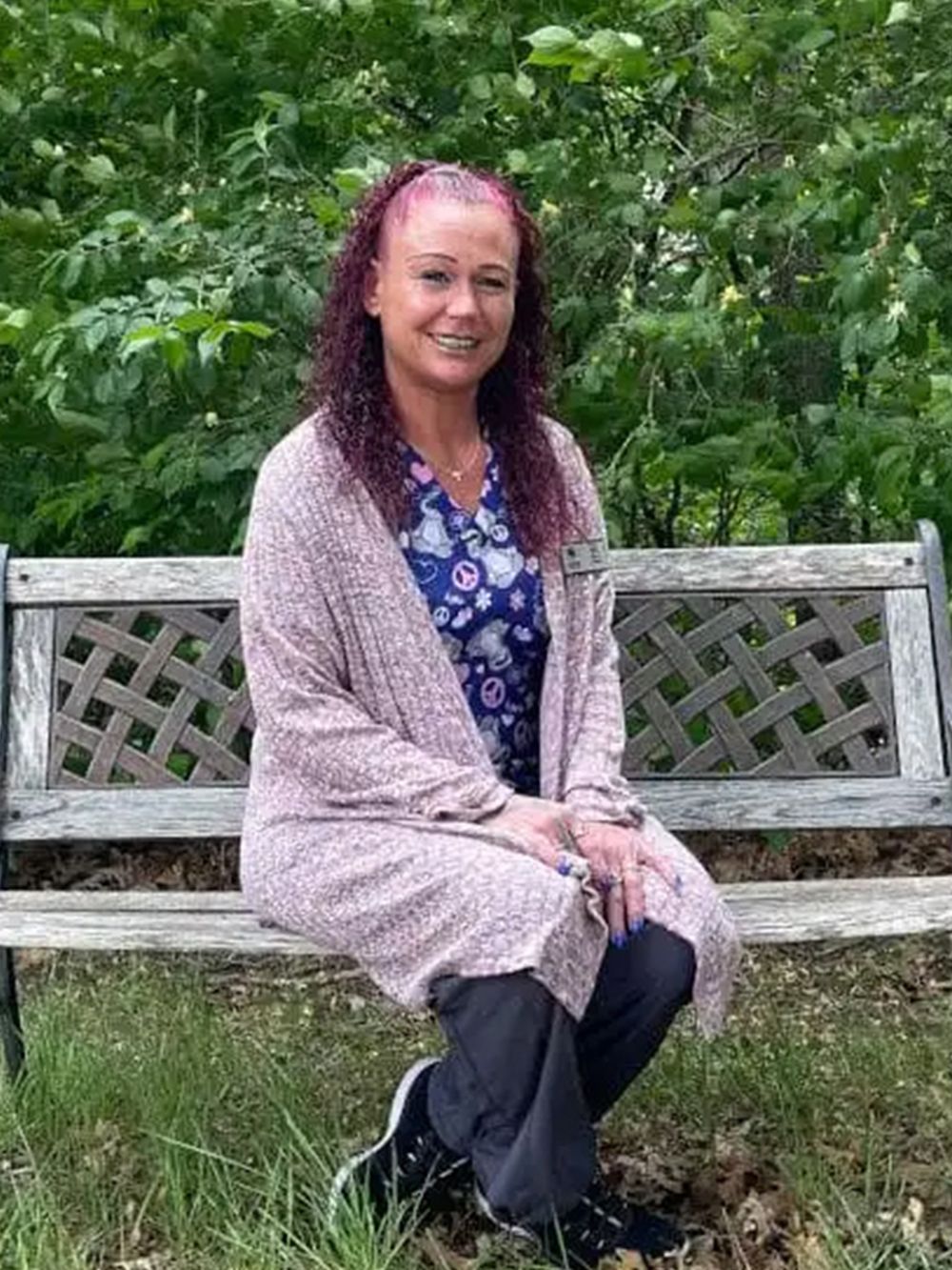 Woman with curly red hair sits on a wooden bench, wearing a pink sweater and scrubs, in a park.