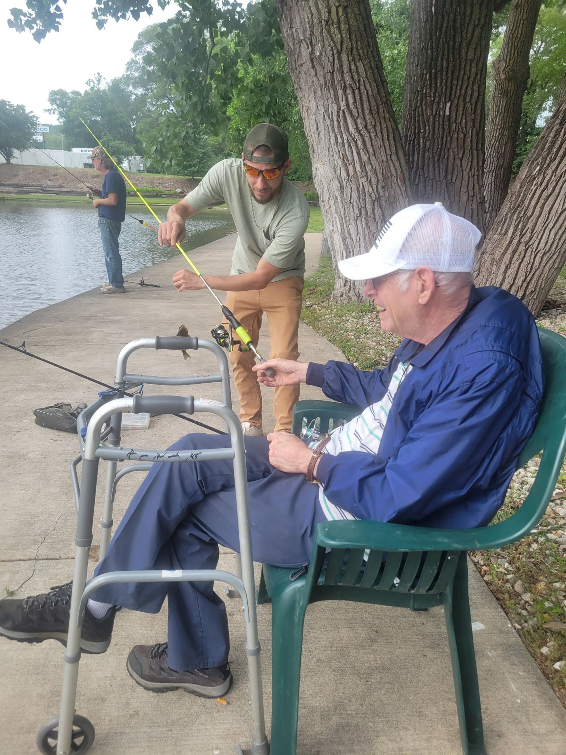 Man in a blue jacket sits in a chair near a pond. Another man helps him with a fishing rod. A third person fishes in the background.