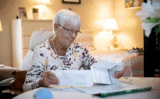 Woman with glasses smiling, examining papers at a table in a well-lit room with a bed and dresser in the background.