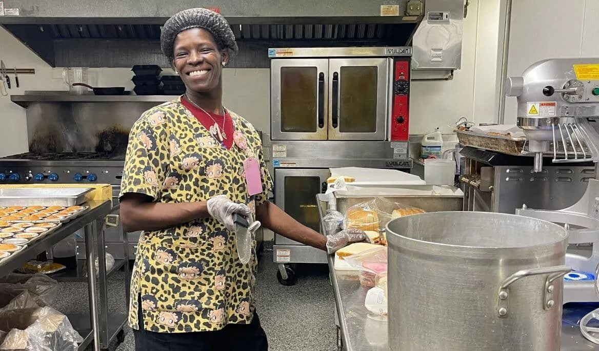 Person smiling in kitchen, wearing a floral uniform. Pots, ovens, and food visible in the background.