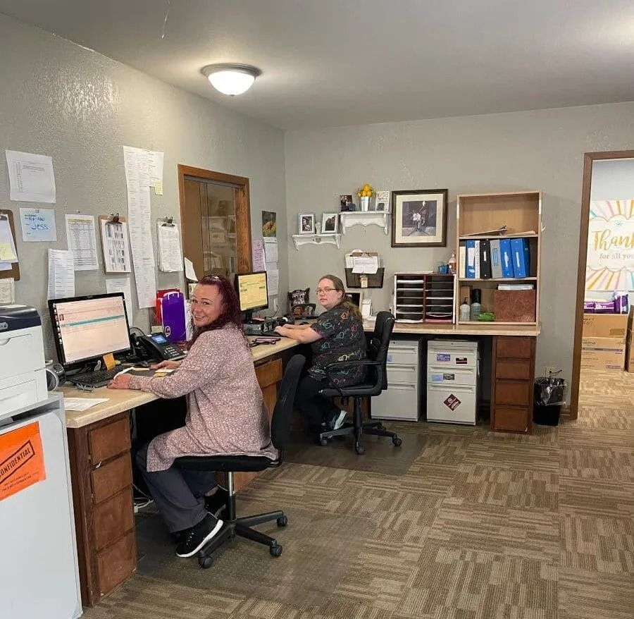Two people work at desks in an office. One smiles, the other looks at a computer. Papers and storage are visible.