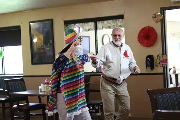 Two people dancing in a restaurant; one in a colorful hat and poncho, the other in a white jacket.