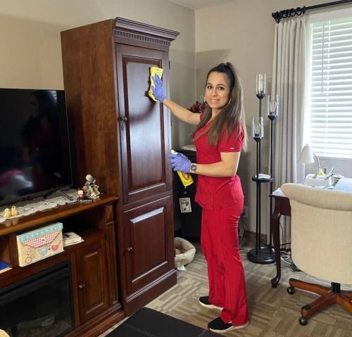 Woman in red scrubs cleaning wooden cabinet with yellow spray bottle.