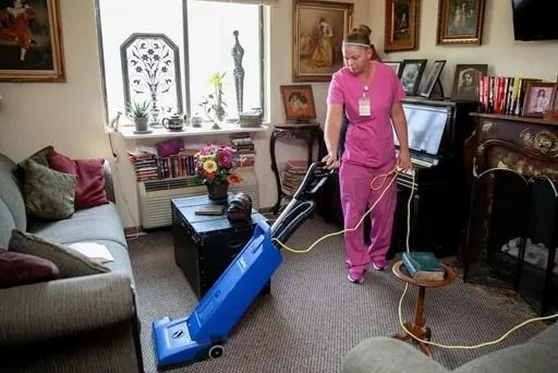 Woman in pink scrubs vacuums a living room with a blue vacuum cleaner.