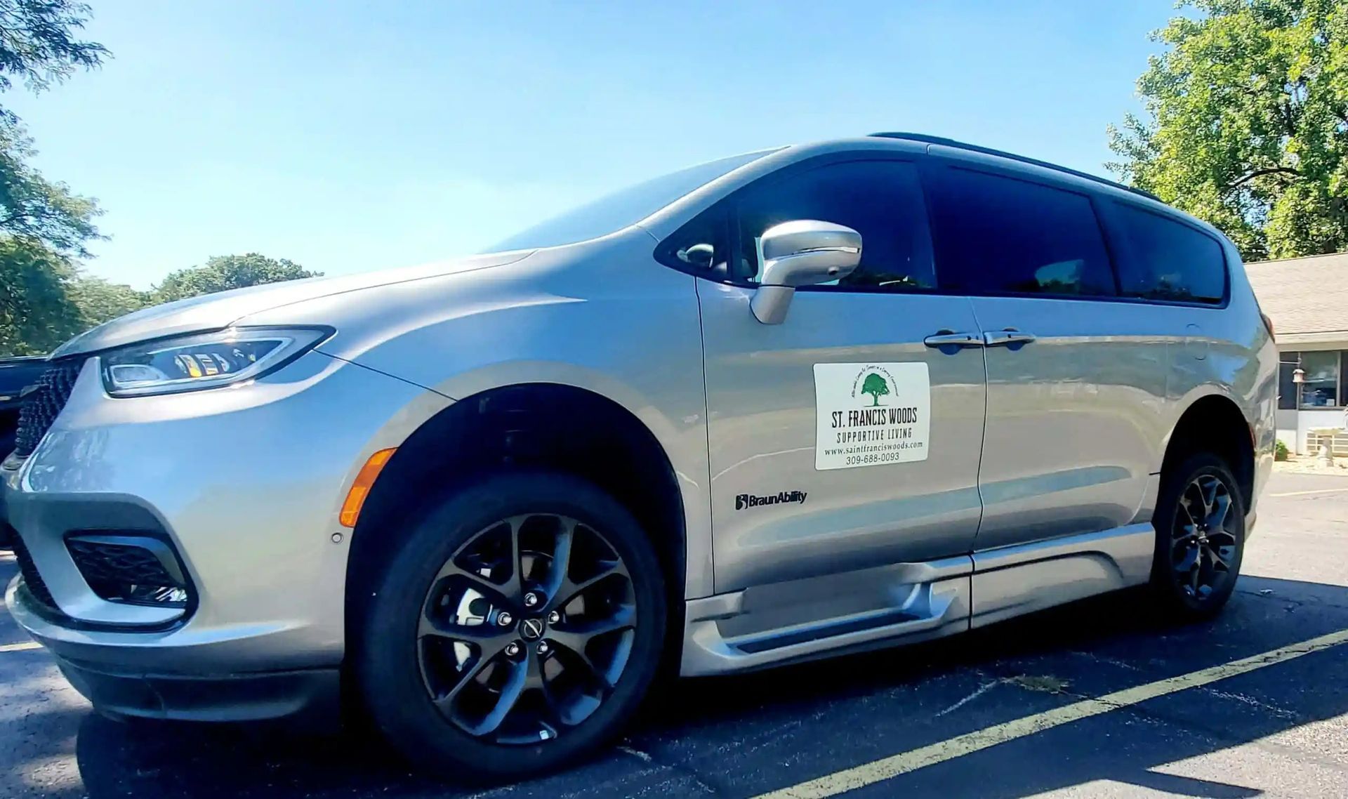 Silver wheelchair-accessible minivan parked outdoors. Has a company logo on the side and a lowered door for entry.