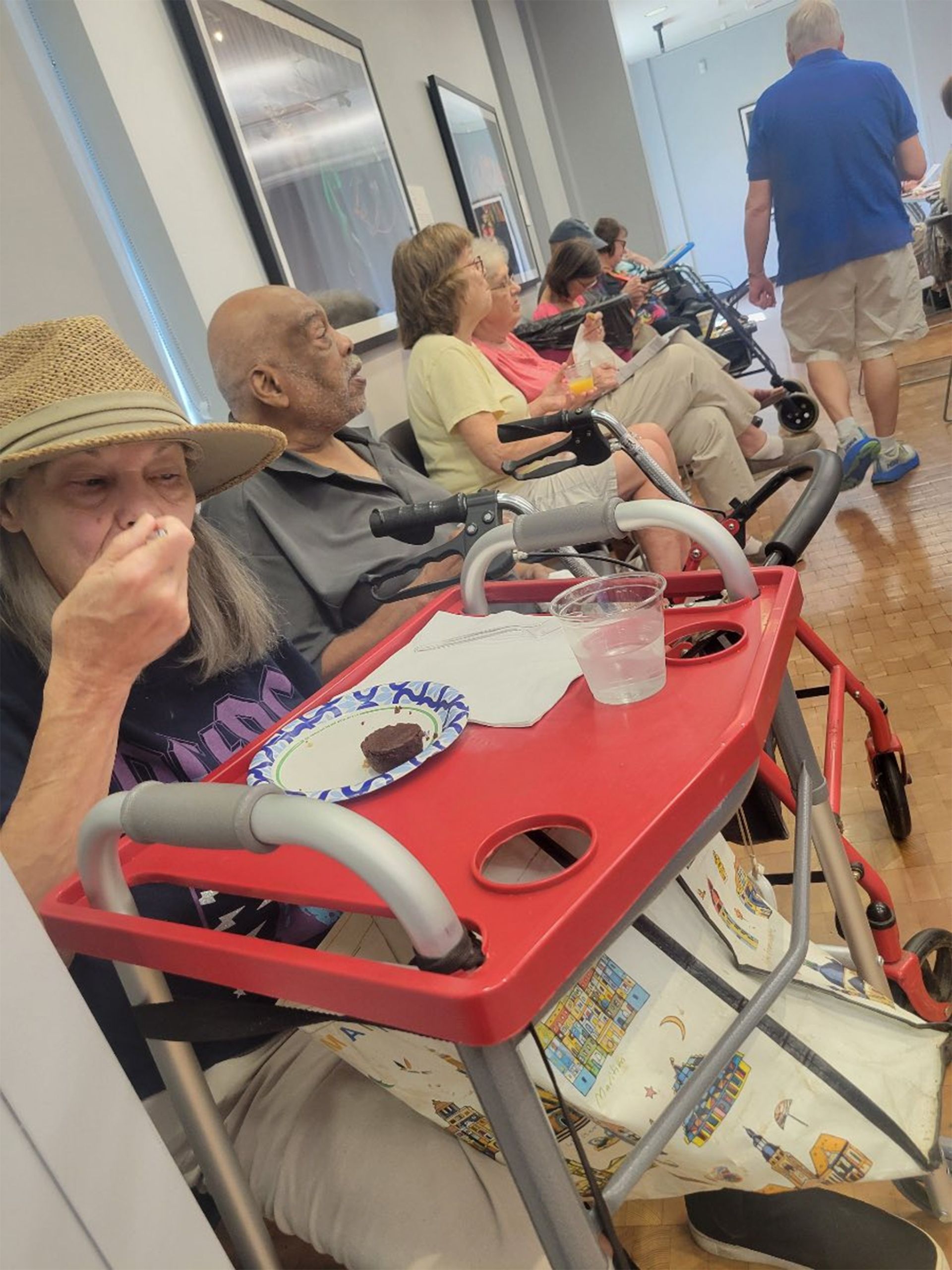 People seated in a room, some using walkers. One person eats from a plate, another walks past.