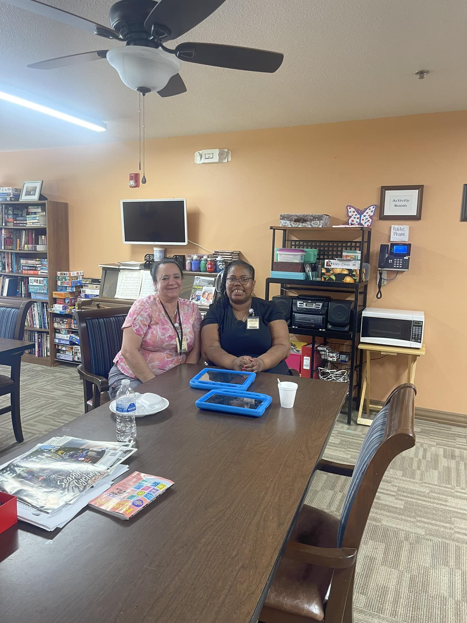Two people seated at a table, smiling at the camera in a room with a TV, microwave, and bookshelf.