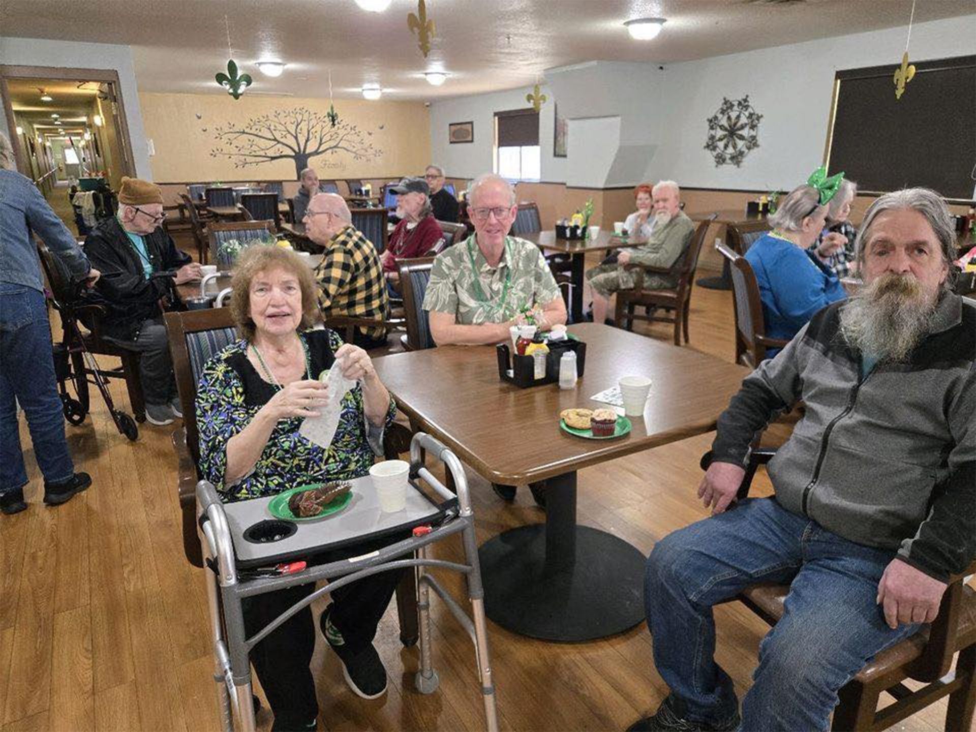 People seated at tables in a dining room. Woman in front has a walker and is holding a drink. Others are eating and talking.