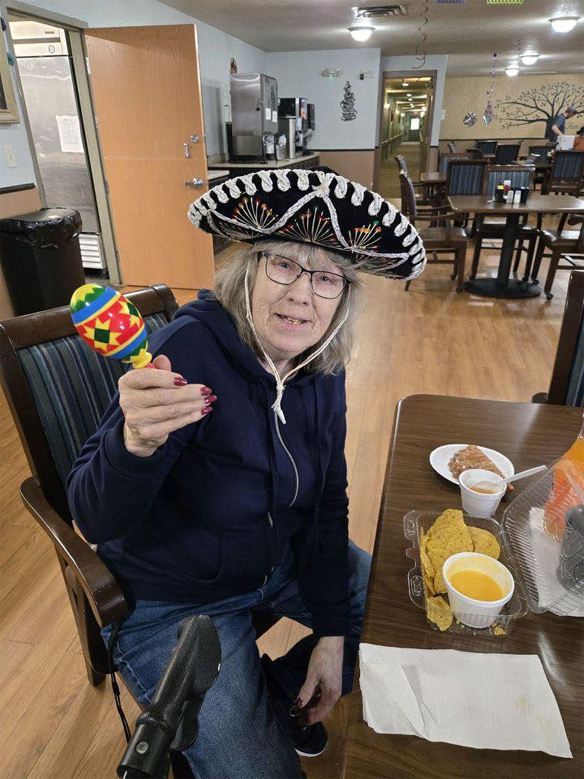 Woman in sombrero holding maraca, sitting at table with chips and cheese.