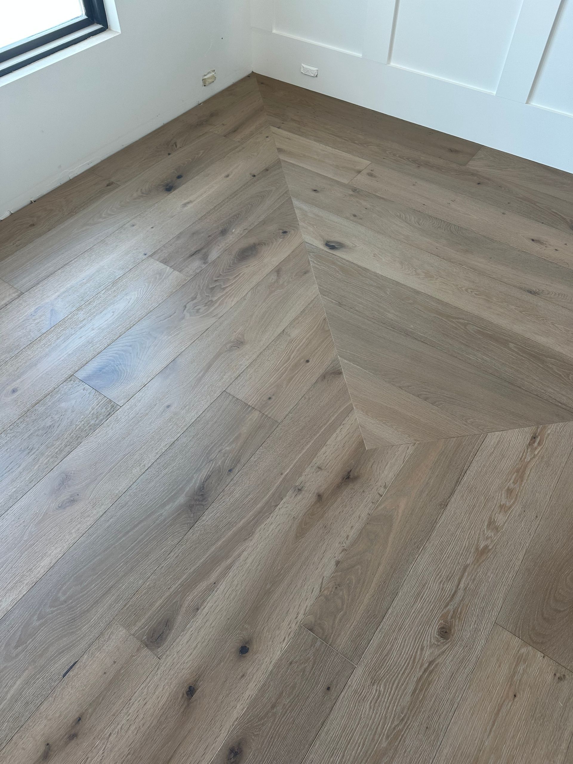 Herringbone wood flooring in a corner, with a light brown color and white wall.