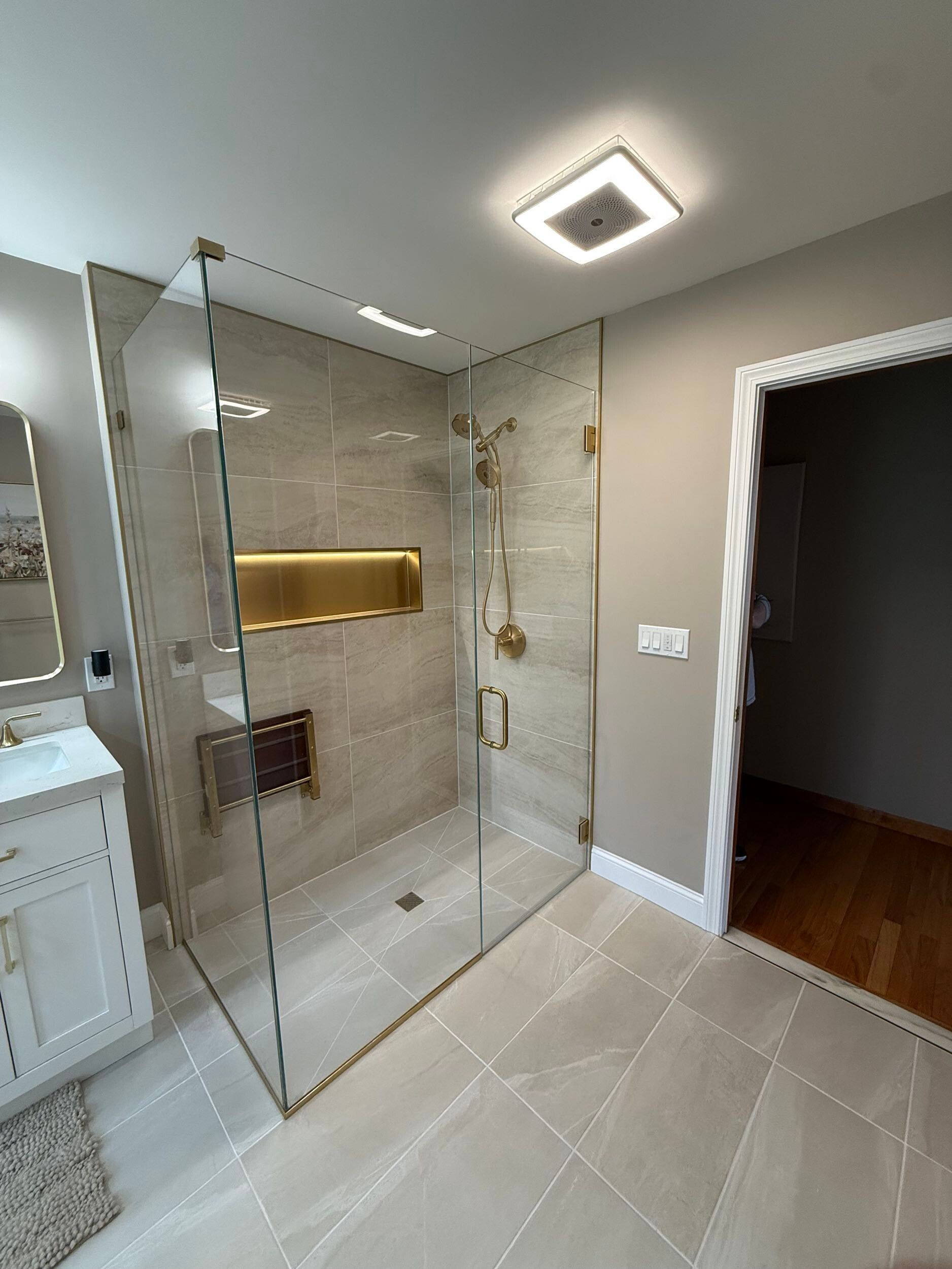 Modern bathroom with glass shower, gold fixtures, beige tile, and open doorway to a dark hall
