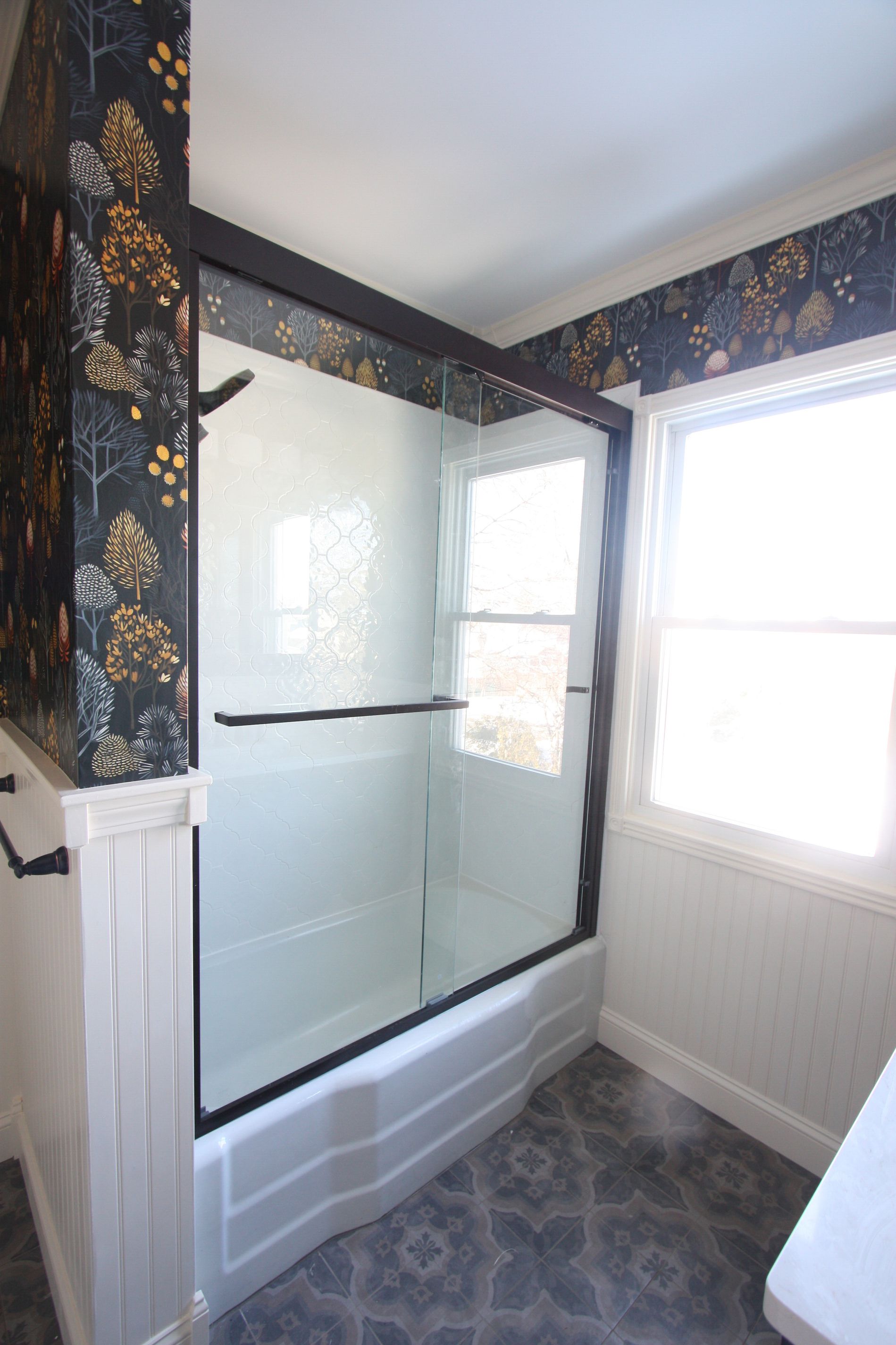 Small bathroom with white tub, glass shower doors, floral wallpaper, and a window.