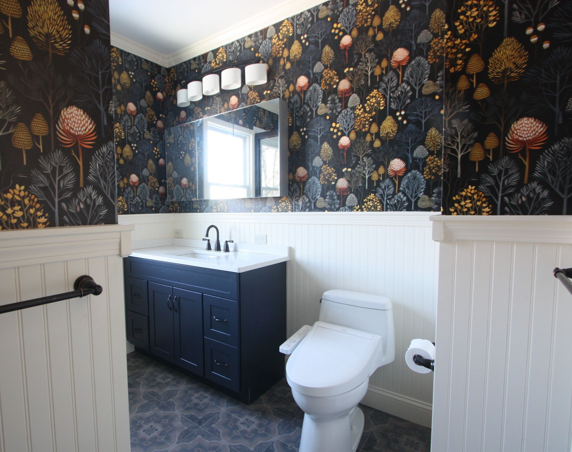 Modern bathroom with floral wallpaper, white wainscoting, dark vanity, toilet, and window near the sink