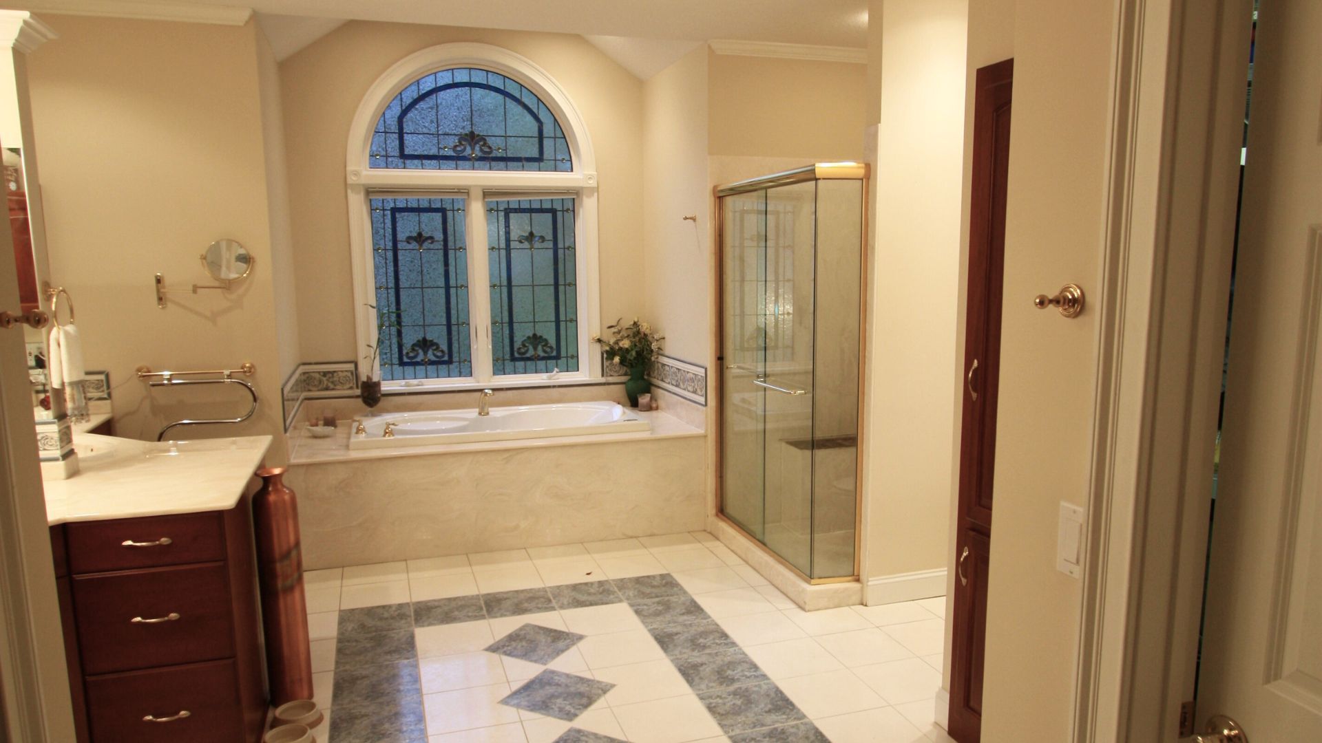 Spacious beige bathroom with arched window, glass shower, vanity, and tiled floor with blue accents
